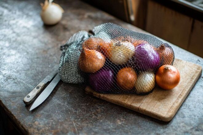Une variété colorée d'oignons dans un sac en filet, posée à côté d'une planche à découper et d'un couteau.